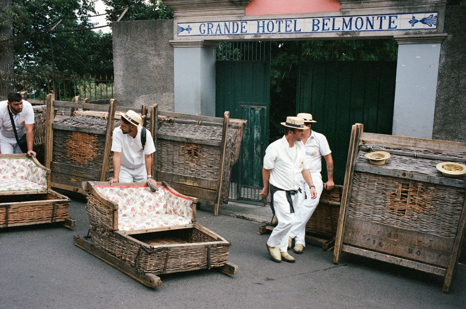 Color film photograph of men in white uniforms moving wicker sleds beneath the Grande Hotel Belmonte sign